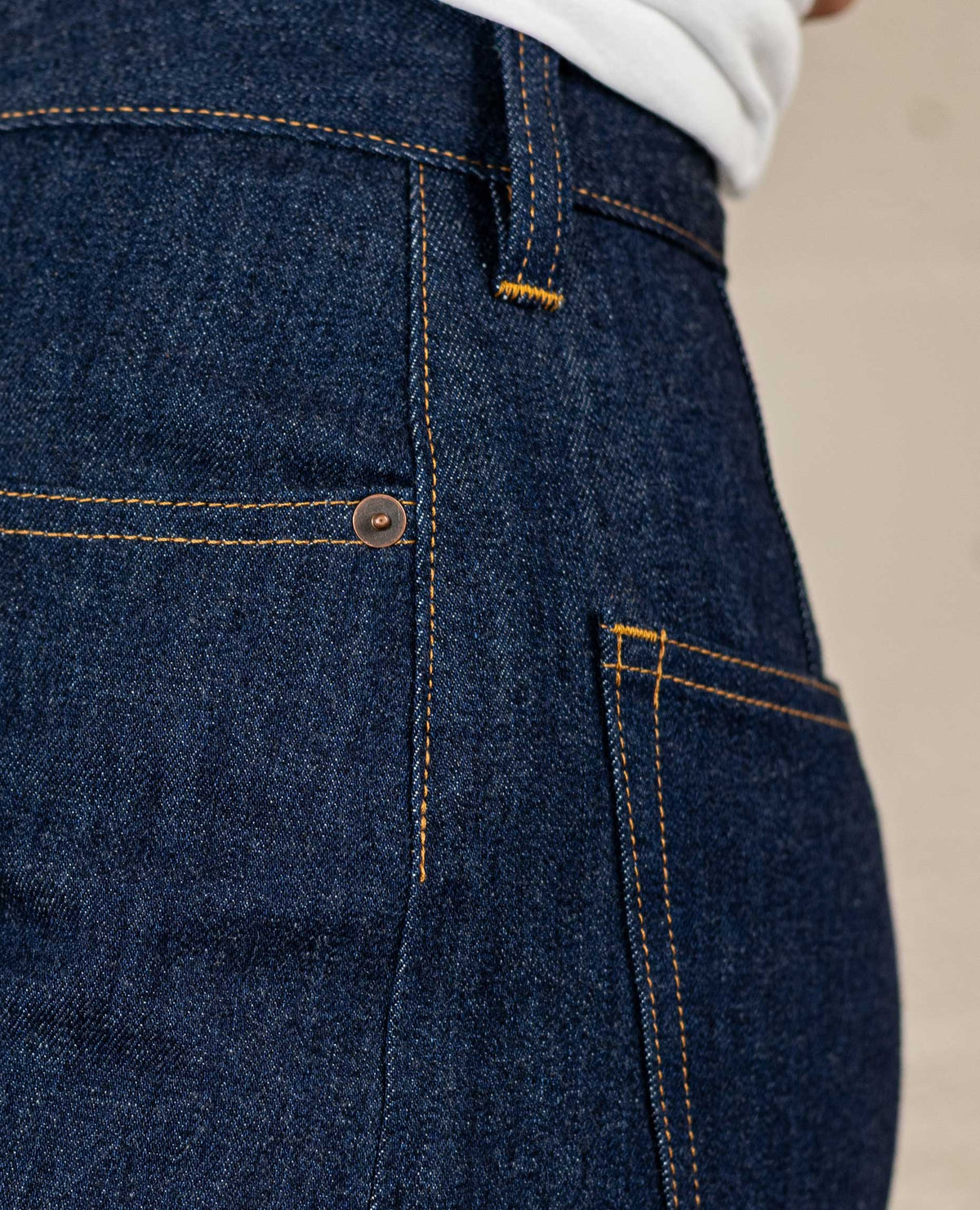 Close-up of dark indigo denim jeans showing the back pocket, pocket rivet, and contrast orange stitching.